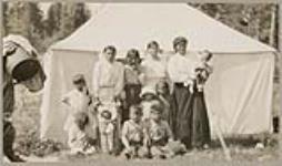 [Group of Anishinaabe women, children, and baby in a tartan cloth tikinagan] 1920