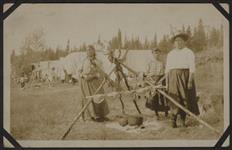 [Three Anishinaabe women standing in front of drying meat over fire during Treaty time, Obishikokaang (Lac Seul First Nation), Ontario 1928] 1928