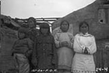 Group of Eskmimos. Nu-kood-lah's wife on right. [Left to right: Akumaluk, Utak, Qillaq, Aarjuaq and Ataguttiaq. They are standing in front of houses built by the R.C.M.P.] 1924