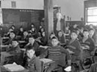 Cree students at their desks with their teacher in a classroom, All Saints Indian Residential School, Lac La Ronge, Saskatchewan, March 1945
 mars 1945