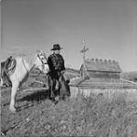 Pan Phillips on his way to the Quesnel, B.C., cattle sale, with his white horse, Queenie, stands at one of the gravehouses in the Kluskus cemetery. This is characteristic of many Indian cemeteries in central B.C Oct. 1956