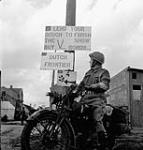 Lance-Corporal J.D. Robillard of The Royal Regiment of Canada passing a sign which reads "Lend Your Dough To Finish The Show - Buy V Bonds", Putte, Netherlands, 14 October 1944 October 14, 1944.