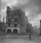 Unidentified member of the Canadian Provost Corps directing traffic past a burning building 17 Aug. 1944