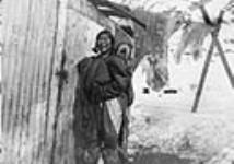 Inuit woman and child outside wood buildings. Skins hanging in background