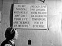 Woman worker reads poster, "DO NOT CONCEAL MISTAKES IT MAY COST YOUR LIFE AND THE LIVES OF OTHERS" at the Cherrier bomb-making plant mai 1941