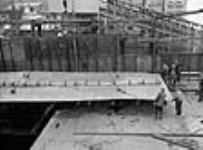 Des ouvriers guident une grue qui descend une grande plaque d'acier sur un navire de charge en construction Sept. 1943