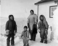 Inuit family at the Roman Catholic Mission. [This photograph, taken by a member of the Roman Catholic church around the month of April, includes from left to right an unidentified boy, Theresie Tungilik, Genova (Kadslajuuqaaq) and Panannaaq Alexina Nanurluk who are not wearing heavy caribou parkas.] 1953