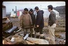 James Houston, Terry Ryan and two men in conversation beside a pile of wood and soapstone, Cape Dorset, Nunavut [between August 24-October 3, 1960].