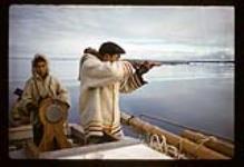 Pitseolak aiming a rifle in a boat steered by his son, Sarpinak, near Iqaluit, Nunavut [between August 16-19, 1960]