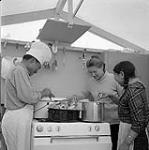 [Barbara Hinds (middle), a man and a woman standing around a stove in a kitchen] [between 1956-1960]