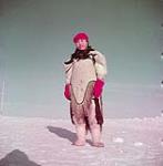 Inuk woman outdoors, wearing a parka, red hat and mittens octobre, 1951.