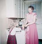 Woman opens a Co-op canned good at kitchen counter while little girl in plaid skirt watches septembre 1953