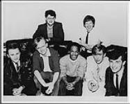 Haircut 100 sitting on a couch following the signing of their record contract with Polydor, London: (back) Trevor Long (band manager), John Briley (Senior A&R Manager, Polydor UK), (front) Graham Jones, Phil Smith, Blair Cunningham, Les Nemes, Mark Fox [entre 1983-1984].