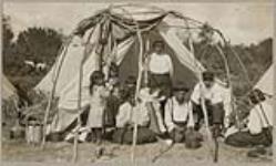 [Group of Anishinaabe men, women, and children underneath a domed wigwam frame] 1920