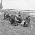 Group of Cree enjoying a game of checkers outdoors with several canoes in the background. 1949