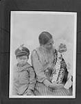 Une femme [Catherine Lafferty] dene assise tient un enfant [Victor Lafferty] dans un porte bébé [Victor Lafferty] avec un garçon à ses côtés, Fort Resolution, Territoires du Nord-Ouest vers 1905-1931.