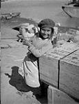 Gwichya Gwich'in girl [Rosa Norman] leaning against a wooden crate by the shore, Tsiigehtchic (Tsiigehtshik, formerly Arctic Red River), Northwest Territories, ca. 1945  c.a. 1945
