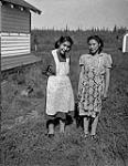 Young Gwichya Gwich'in women [Rose Clark (née Cardinal) and Martha Bullock (née Andre)] wearing floral aprons standing next to a building, Tsiigehtchic (Tsiigehtshik, formerly Arctic Red River), Northwest Territories, ca. 1945  c.a. 1945.