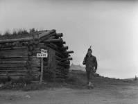 Private Elbert Pieper of the U.S. Army stands sentry duty beside a trapper's cabin containing construction supplies for the Alaska Highway. 1942