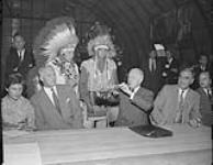 Hon. Lionel Chevrier, Chairman of the St. Lawrence Seaway Authority, smoking a pipe of peace with residents of Caughnawaga. 25 July 1956