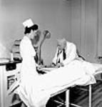 L'infirmière Lora Mitchell prend le pouls d'une ouvrière pendant que le Dr L.E. Mylks effectue une analyse sanguine à l'hôpital de l'usine de la Société Polymer Limitée. juin 1943