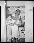Michael Berens' (the photographer) wife Mili with Nellie Lutcher in the "kitchen" of their suite at the Standish Hotel.  Mili fed many entertainers after their performances, after the hotel's kitchen had closed for the night.  Berens writes of Mili's kitchen: "it was the smallest kitchen imaginable, built in a clothes-cupboard with the smallest refrigerator, a two-hole burner, a bread-box, four shelves and a lot of Mount Royal, canned Dainty Rice.". ca. 1950.