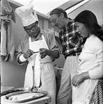 [Mackenzie Porter (middle) and a woman observing a man preparing fish, Iqaluit, Nunavut]. [between 1956-1960]