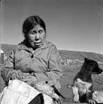 [Woman sewing while seated outside next to a dog, Iqaluit, Nunavut]. [between 1956-1960]