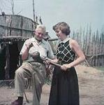 Woman holding wooden implement speaks to elderly man, Midland. August 1955