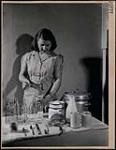 Jeune femme vêtue d'une robe rayée remplissant un biberon de lait. Illustration pour la publication du gouvernement « Canadian Mother and Child (La mère canadienne et son enfant) ». mars 1946