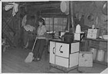 Aboriginal girl sweeping floor of log house beside stove. n.d.