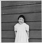 Fille souriante portant une robe blanche, debout contre un mur, à l'extérieur. Hôpital Charles Camsell, Edmonton (Alberta) juillet 1958