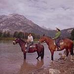 [A man and a woman on horseback at the edge of a river, Alberta]  August 1956