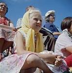 Children and adults are interested onlookers of the activities on the outdoor stage at the Polo Park, Winnipeg, Manitoba during Dominion Day celebrations.  01 juillet 1961