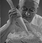 Un chef place le glaçage sur un gâteau à l'aide d'une poche et d'une douille. 1957