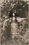 [Jessee (or Jim) Snow standing in the bush, wearing fringed dress, headband and feather]  1912.