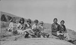 Group of Métis children and two women sitting on a large rock, Fort Chipewyan, Alberta  vers 1930-1939.