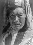 A Haida of Massett, British Columbia. [Identified as Hayas of Kayung.] The head-dress is a "dancing hat", and consists of a carved wooden mask, surmounted by numerous sea-lion bristles and with pendent strips of ermine-skin 1916