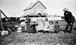 Group of children of varying ages with a man standing on grass in front of a frame house, Île-à-la-Crosse, Saskatchewan ca. 1905-1931.