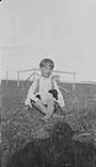 Garçon assis dans le gazon avec un chiot sur ses genoux, Île à la Crosse (Saskatchewan) ca. 1905-1931.