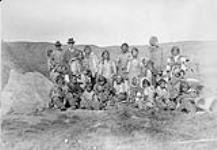 Group of Inuit men, women and children and two non-Inuit men standing and seated outdoors at Kuujjuarapik (formerly Whale River), Quebec