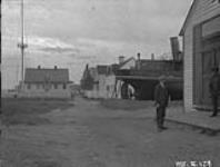 A Cree man and a non-Indigenous man standing in front of buildings and a boat August 1926.