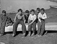 Gwichyaa Gwich'in boys [(left to right) JP Kendo, unidentified, Billy Cardinal, Cecil Andre, William Clark, John Norbert, and Victor Simon Modeste] sitting on a canoe, Tsiigehtchic (Tsiigehtshik, formerly Arctic Red River), Northwest Territories, ca. 1945 c.a. 1945