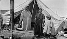 Two Gwichya Gwich'in women, one making bannock, and a girl standing outside of a tent 1930
