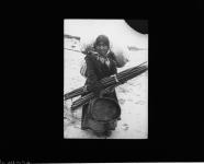 A Cree woman carrying a basket and wooden poles, possibly in Chisasibi (formerly Fort George), Quebec 1910-1922.