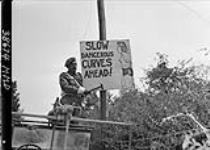 Sign indicating " Slow, Dangerous Curves Ahead " being put up along the road of Falaise by Cpl. Ernie Nault, Meadow Lake, Sask., Canadian Provost Company 14 Aug. 1944