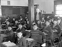 Élèves cris à leurs pupitres avec leur professeur dans une salle de classe, Pensionnat indien de All Saints, Lac La Ronge (Saskatchewan), mars 1945  March 1945.