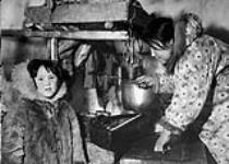 [Inuit woman and young girl named Marian Bolt (Hayohok) tending the seal oil lamp inside an igloo] Original title: Same youngster inside igloo with her mother who is tending the seal-oil lamp which provides warmth and light for the snowhouse 1949.
