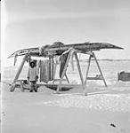 Inuit man standing near racks where kayaks are kept during winter Jan. 1946