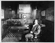 W. Howard, radio operator of radio station VAK seated at desk with radio equipment on it 1912